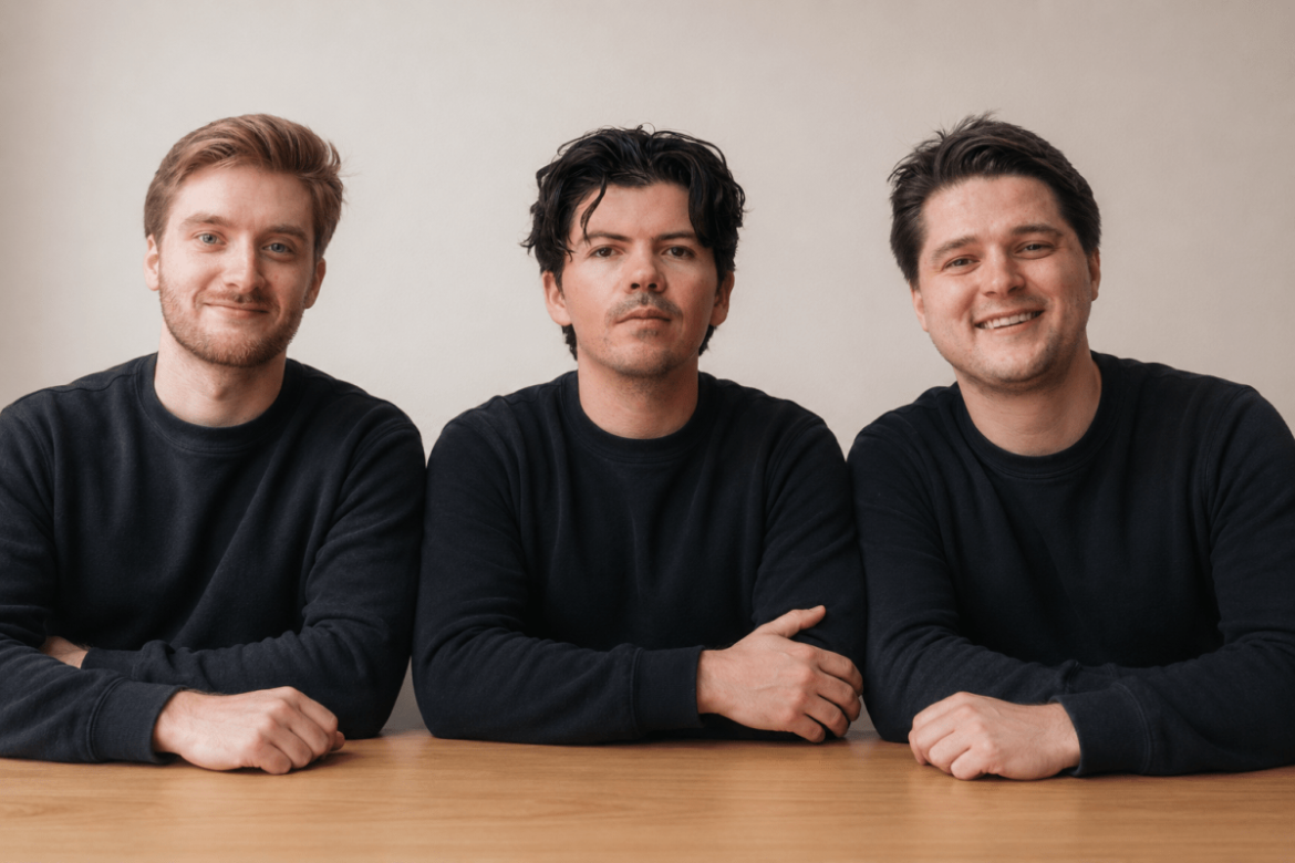 Three caucasian men in black long-sleeved crewneck shirts sit at a desk against a beige background.